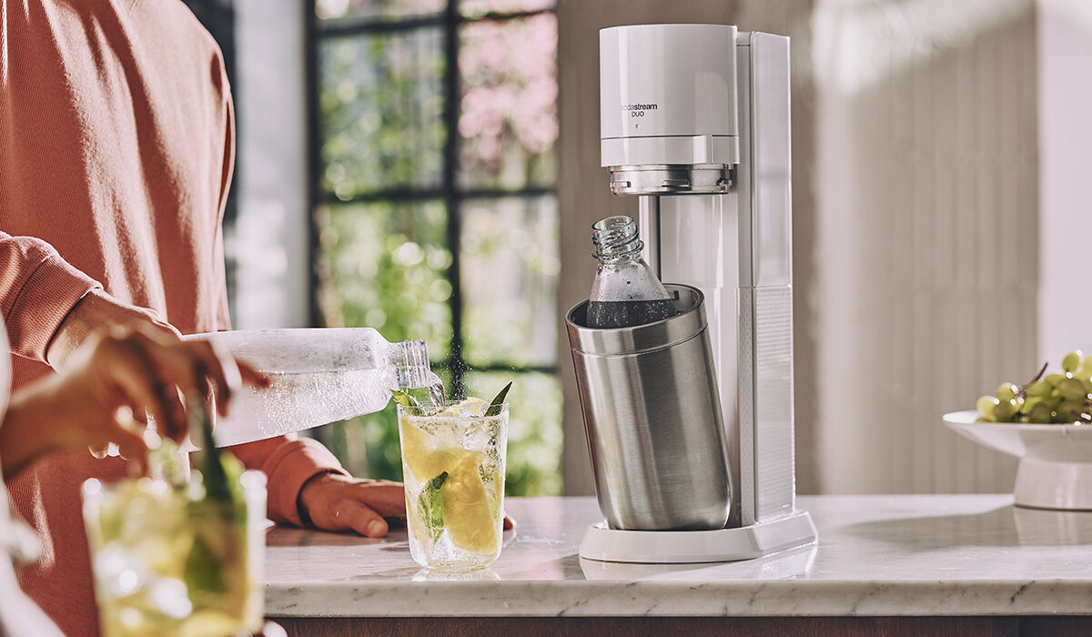 Ekspres SodaStream Duo Biały widok przygotowywanie gotowego napoju przez kobietę i urządzenie na tle kuchni