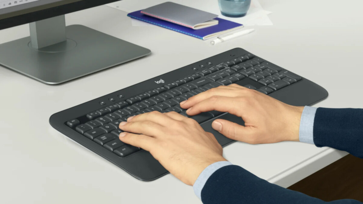 Klawiatura i mysz Logitech MK540 Advanced, grafika przedstawia ręce na klawiaturze leżącej na biurku, w tle monitor, notatniki, długopis oraz kubek
