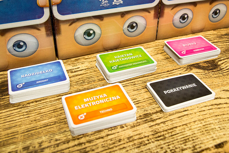 Gra karciana Portal Games Czółko 2-8 graczy grafika przedstawia trzy opakowania gry oraz karty ułożone w stosy na stole