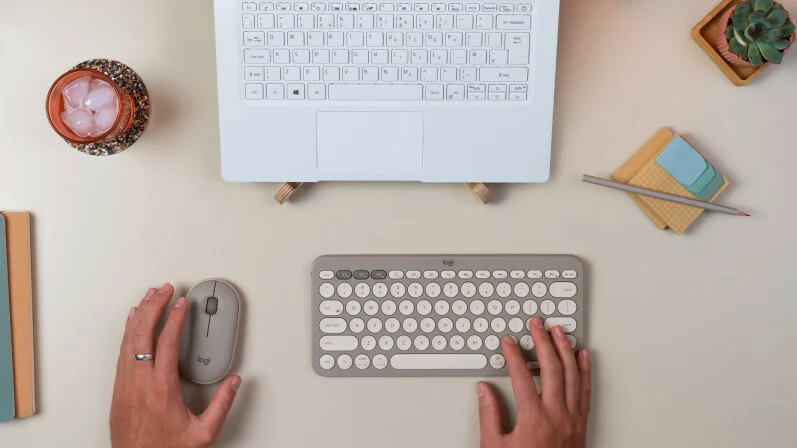 Mysz Logitech Pebble M350 w dłoni mężczyzny wraz z klawiaturą i laptopem w tle