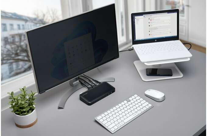 Kabel blokady do laptopa Kensington NanoSaver czarny podłączony do laptopa stojącego na podstawce, pod nią znajduje się telefon, który stoi na biurku obok monitora, routera, klawiatury, myszki oraz kwiatu, w tle widać okno z budynkiem.