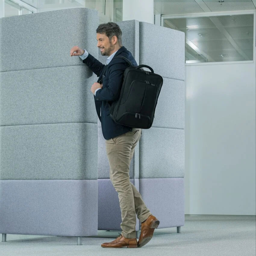 Plecak Dicota Eco Backpack Pro czarny mężczyzna stojący w budynku z plecakiem na ramieniu opierający się o ścianę