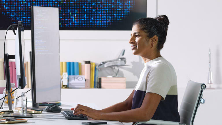 Klawiatura Logitech Mx Mechanical szaro-czarna na biurku, przy którym siedzi kobieta klikająca na klawiaturze przed monitorami w tle książki i obraz mozajka niebieska