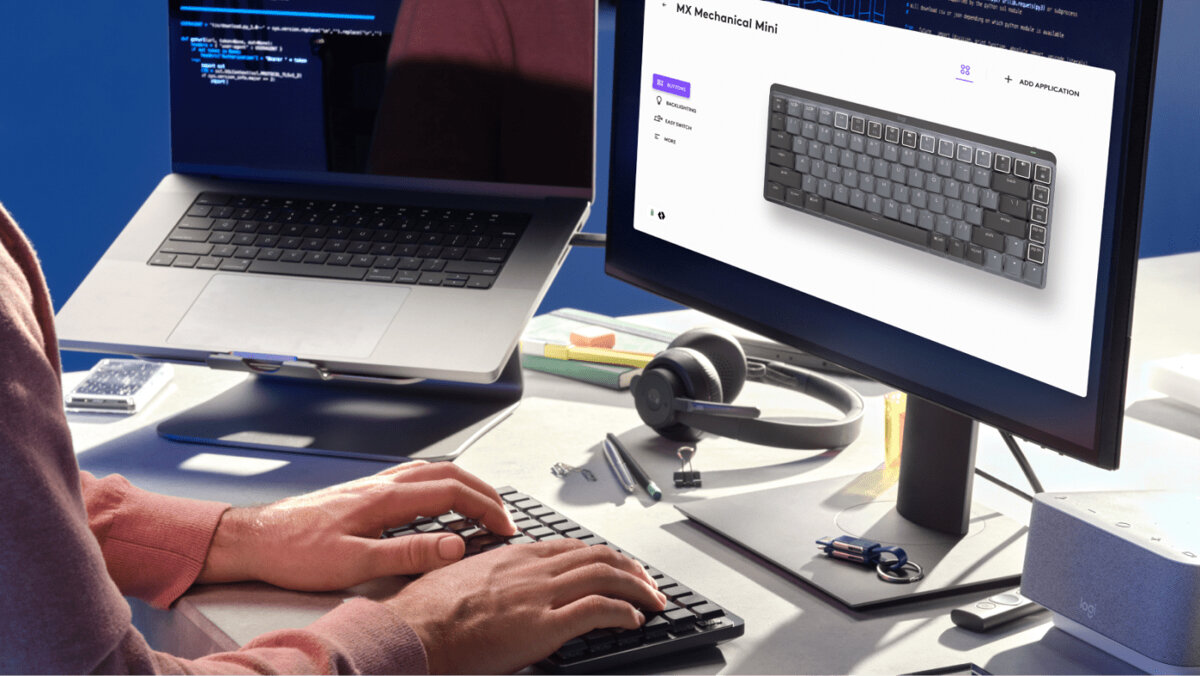Klawiatura Logitech Mx Mechanical szaro-czarna na biurku z klikającymi rękoma mężczyzny, właczony monitor oraz laptop, słuchawki i inne przedmioty na biurku