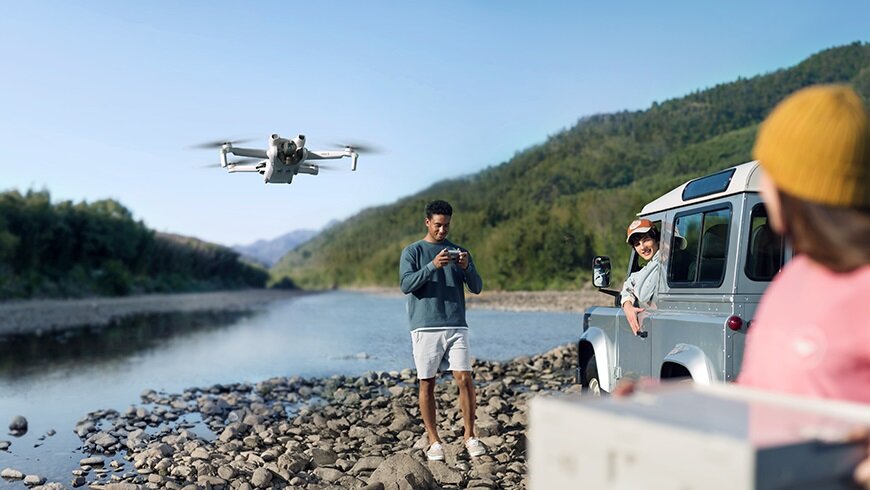 Dron DJI Mini 3 Fly More Combo CP.MA.00000610.01 widok na mężczyznę siedzącego w samochodzie oraz na mężczyznę stojącego na kamieniach w trakcie sterowania latającym nad nim dronem