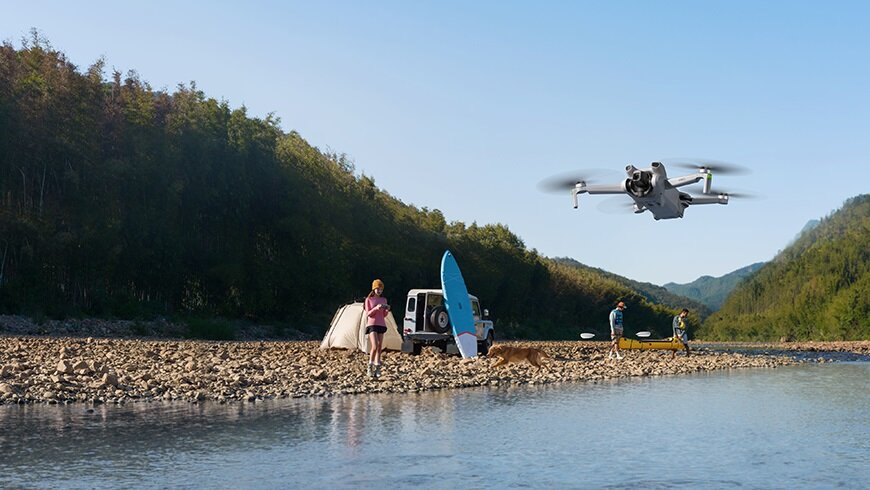 Dron DJI Mini 3 Fly More Combo CP.MA.00000610.01 latający nad wodą oraz sterująca nim kobieta stojąca na kamieniach przy brzegu
