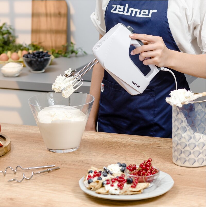 Mikser ręczny Zelmer ZHM2550 biały w ręku kobiety wraz z dodatkowymi mieszadłami oraz owocami na talerzu i pojemnikiem z produktami obok na stole, w tle na szafce produkty spożywcze