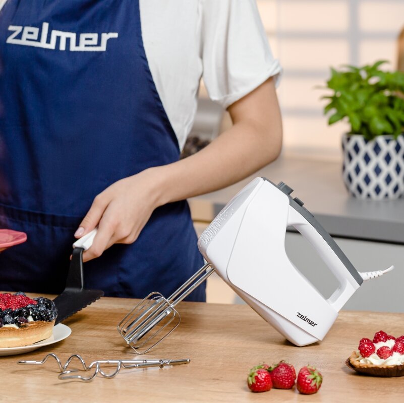 Mikser ręczny Zelmer ZHM2550 biały od lewego boku na stole obok dodatkowe mieszadła, kobieta wkładająca łopatką ciasto, w tle kwiatek