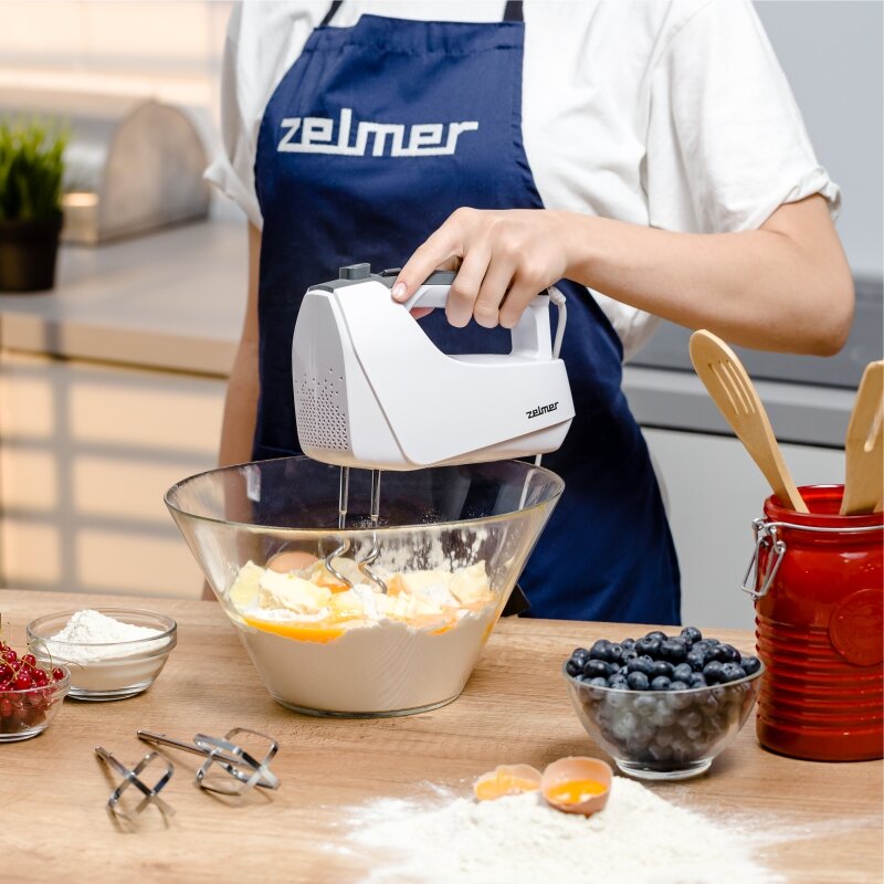 Mikser ręczny Zelmer ZHM2550 biały kobieta mieszająca mikserem masę w misce wraz z wypełnionymi obok miskami na stole