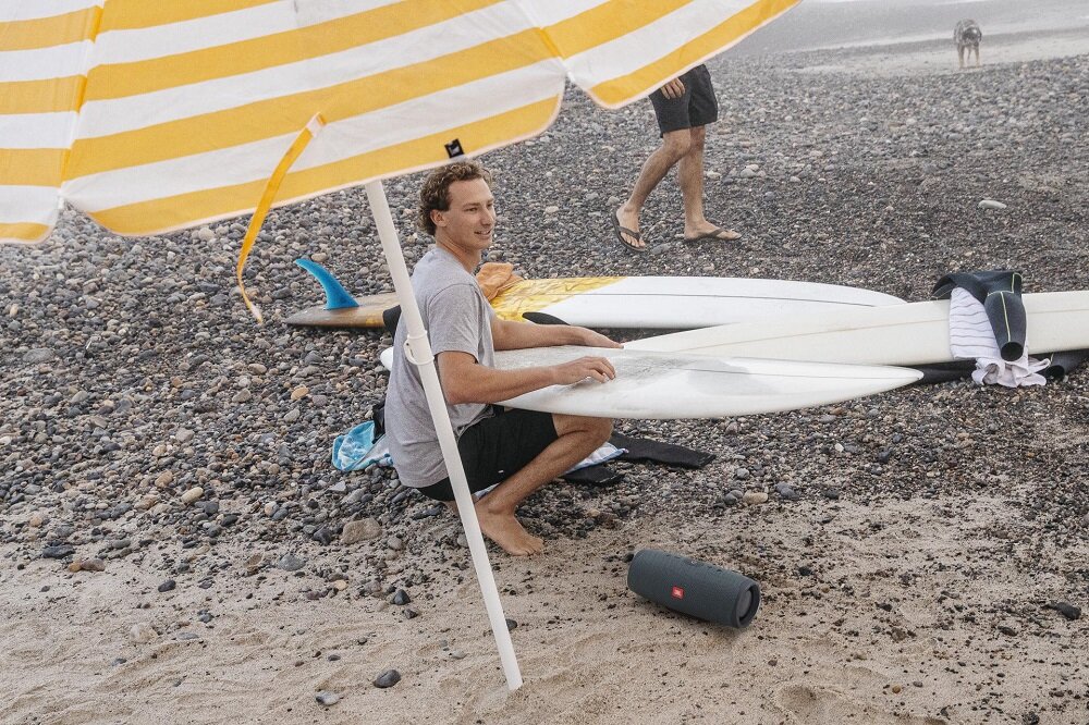 Głośnik JBL Charge Essential 2 widok na mężczyznę trzymającego na kolanach deskę surfingową oraz na głośnik leżący pod skosem na piasku