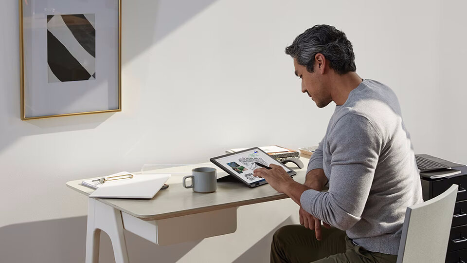 Tablet Microsoft Surface Go 2 4/64GB grafika przedstawiająca mężczyznę siedzącego przy biurku na którym znajduje sie tablet, kubek oraz dokumenty, w tle na ścianie wisi obraz, a za mężczyzną stoi szafka na której leży laptop