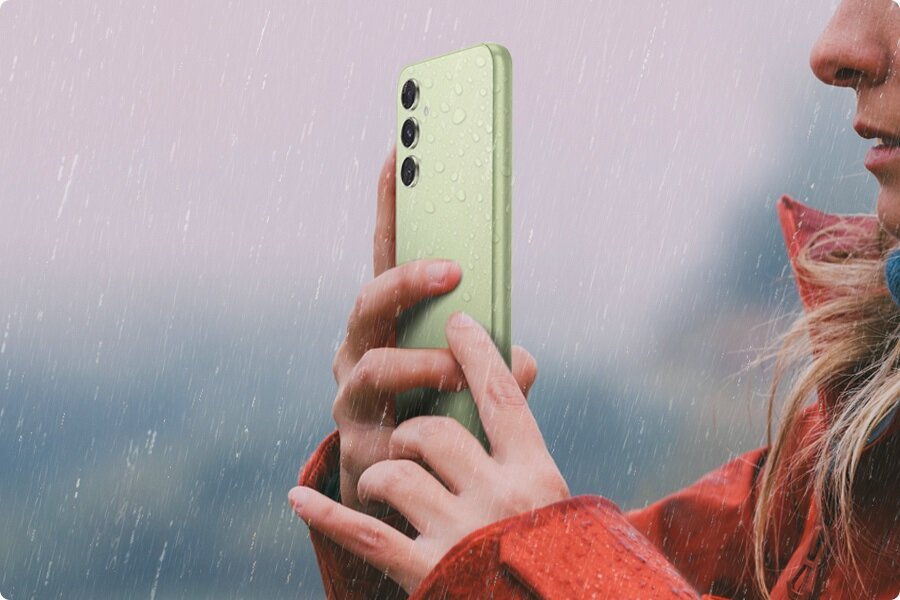 Smartfon Samsung Galaxy A54 5G SM-A546B SM-A546BLVCEUE widok na plecki smartfona trzymanego w dłoniach z kroplami deszczu widocznymi na nim