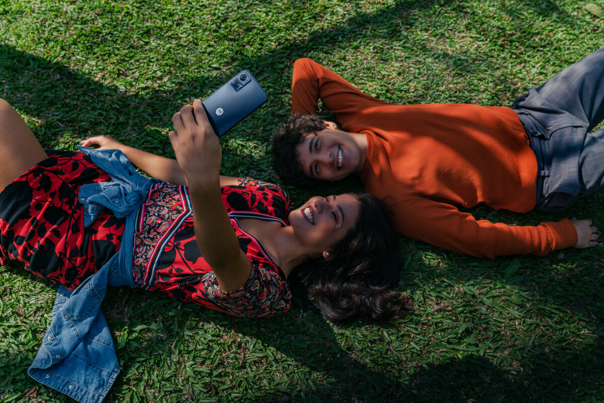 Smartfon Motorola moto e22 od tyłu, w dłoni kobiety robiącej zdjęcie sobie i chłopakowi leżącym na trawie