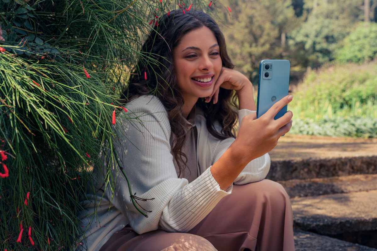 Smartfon Motorola moto e22 od tyłu, w dłoni kobiety siedzącej w ogrodzie i spoglądającej w telefon
