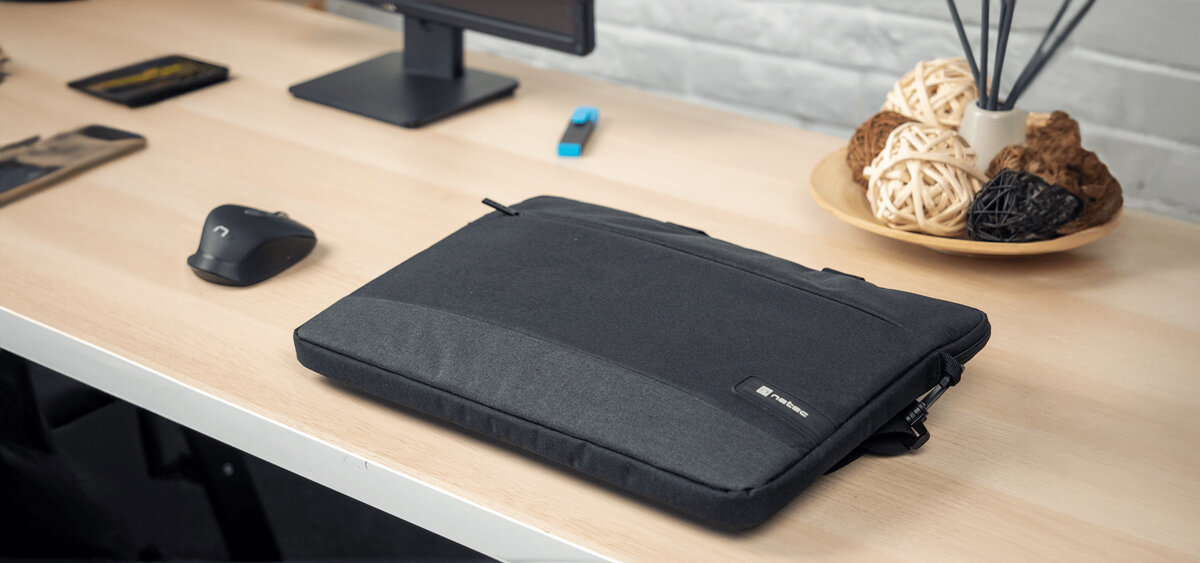 Torba na laptopa Natec Goa 15.6 czarna grafika przedstawia torbę leżącą na biurku obok myszki, w tle widoczna jest dekoracja, mazak, monitor i telefony