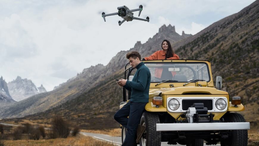 Dron DJI Air 3 Fly More Combo (DJI RC 2) 6000m pokazany mężczyzna sterujący dronem, który unosi się w powietrzu oraz kobieta w samochodzie