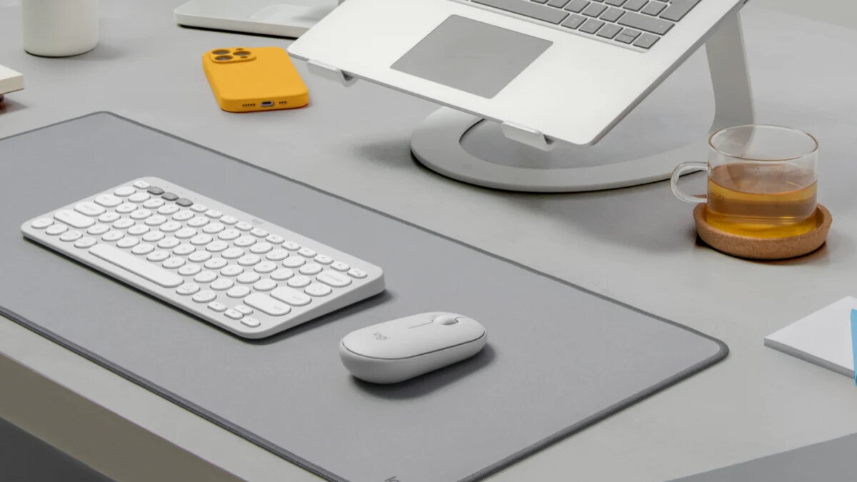 Klawiatura Logitech Pebble Keys 2 K380s biała grafika przedstawia po skosie klawiaturę i mysz leżące na podkładce przed laptopem oraz smartfon