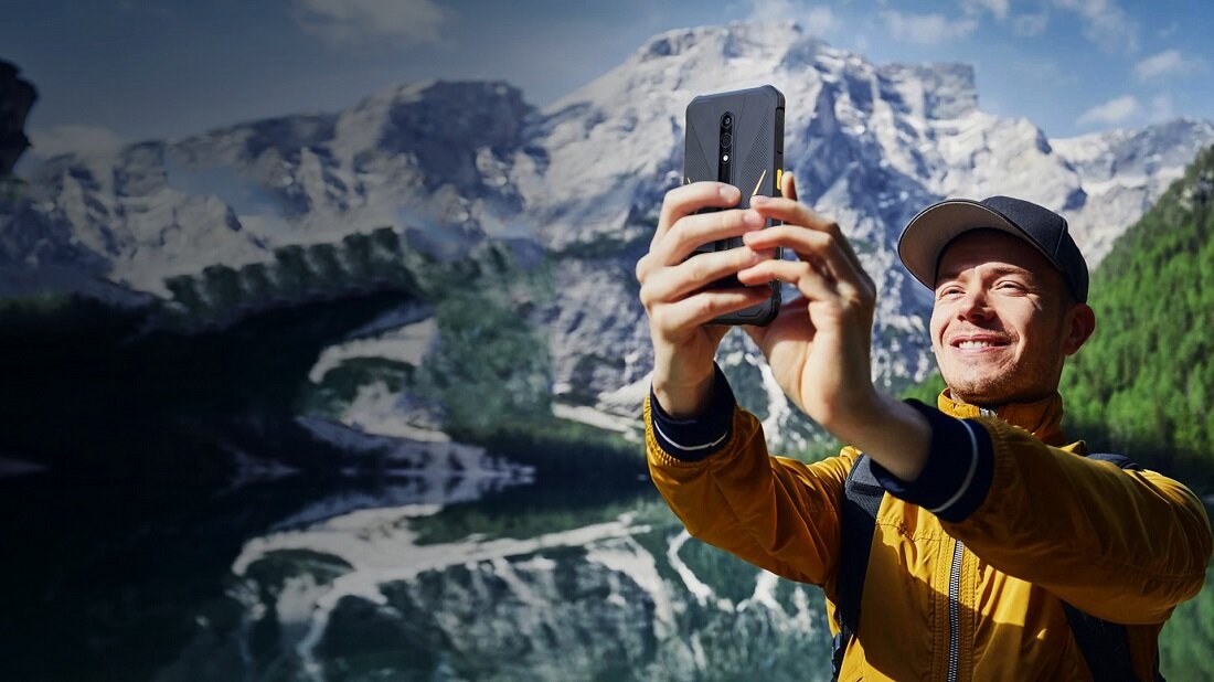Smartfon Ulefone Armor X12 Pro 4GB/64GB czarno-pomarańczowy grafika przedstawia mężczyznę robiącego zdjęcie telefonem w górach